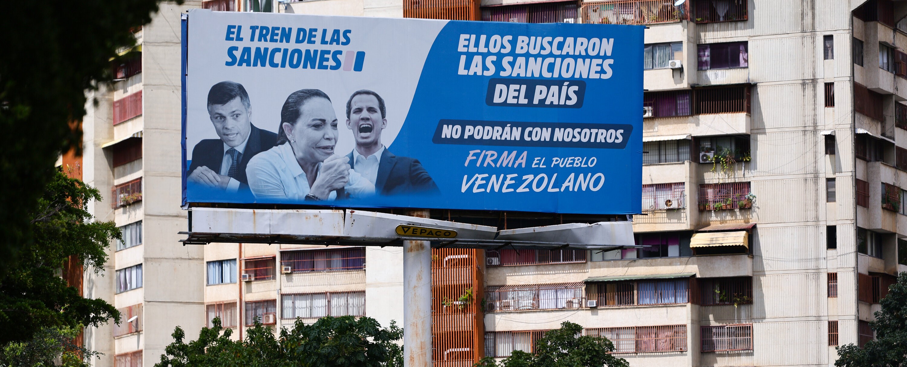 Street of Caracas, October 2025. The poster titled “The Train of Sanctions”, refers to the call for sanctions against Venezuela by the three right-wing leaders in the photo, among them Maria Corina Machado. Implicitly the poster is mocking with President Trump’s accusations against Venezuela of leading the “Train of Aragua” (a criminal gang). The poster text continues: “They will never win over us. The Venezuelan People”. Photo: Jesus Vargas/Getty Images